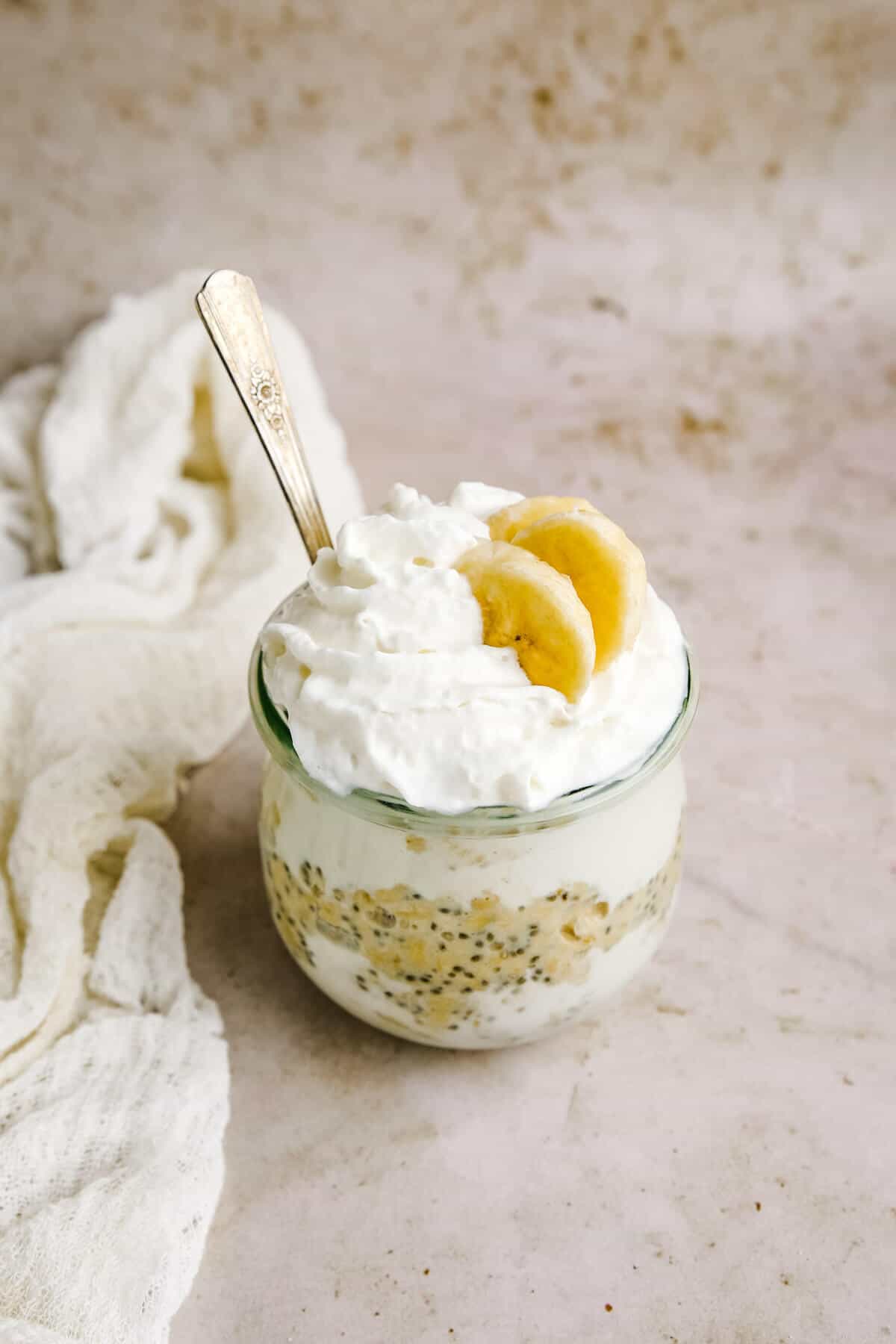 a glass jar filled with layers of protein overnight oats and topped with whipped cream and two slices of banana