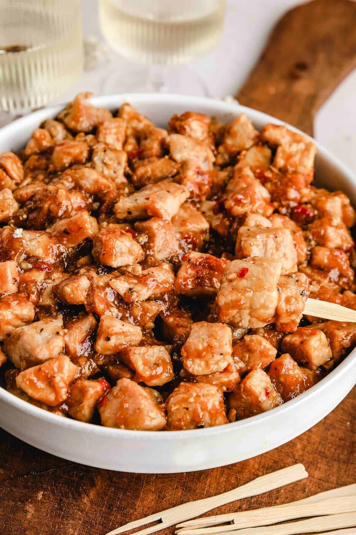 a closeup shot of garlic honey pork bites in a white bowl with one toothpick skewered through three cubed pieces