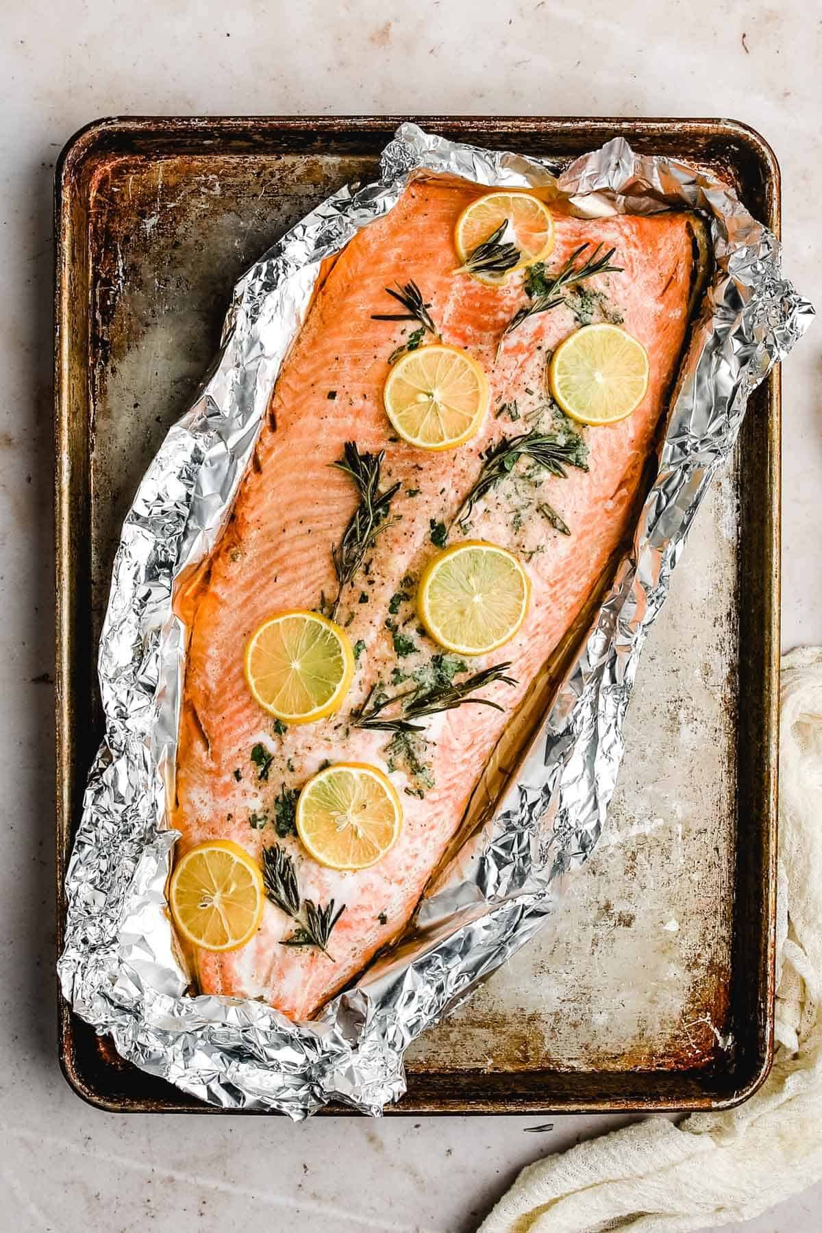 an over head view of a baking sheet with a whole salmon fillet wrapped in foil with fresh rosemary and lemon slices on top