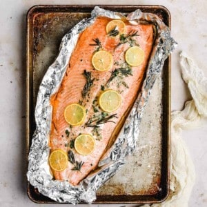 an over head view of a baking sheet with a whole salmon fillet wrapped in foil with fresh rosemary and lemon slices on top