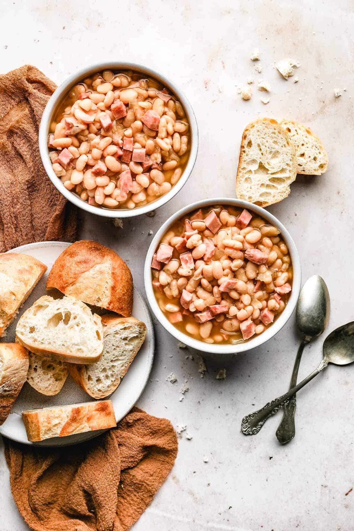 two white bowls filled with instant pot ham and beans next to a white plate of sliced baguette with two silver spoons.