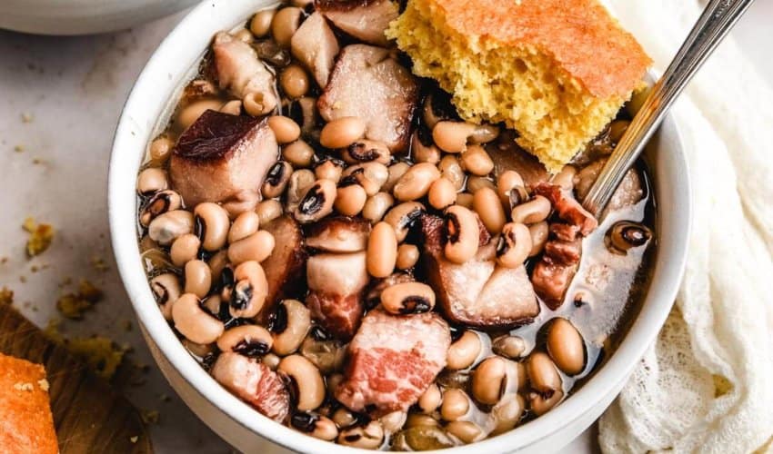 a white bowl filled with hog jowl and black eyed peas with a silver spoon and a slice of cornbread
