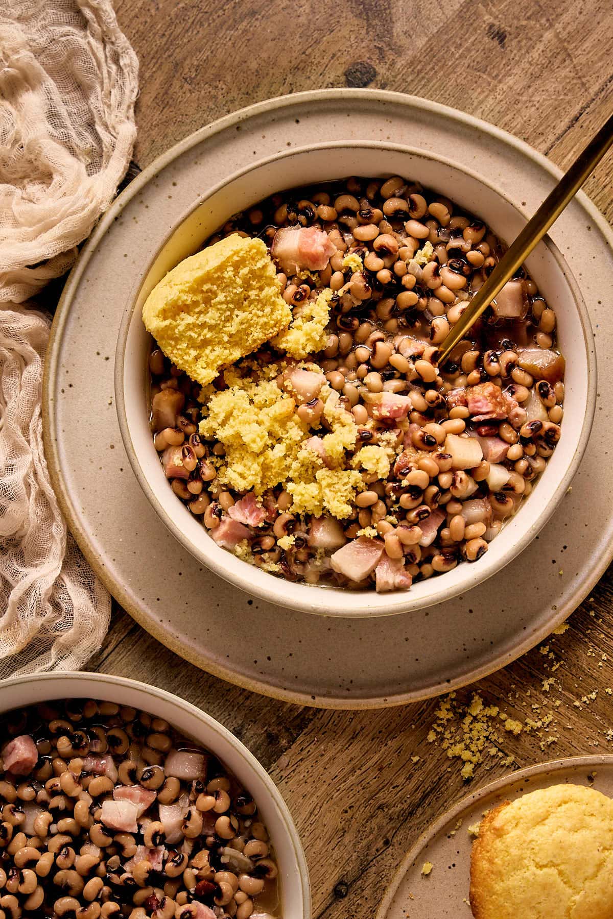 a closeup shot of a cream bowl of pork jowl with southern black eyed peas with a gold spoon and a slice of cornbread. 