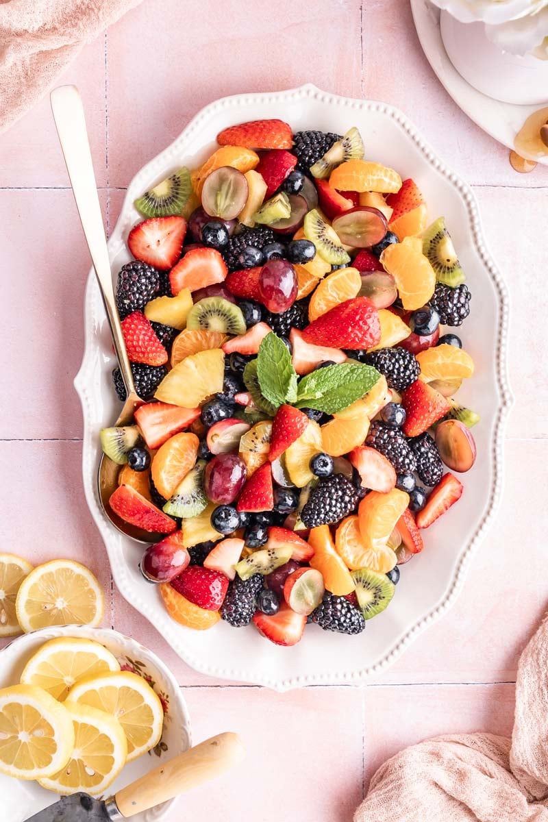 a large white dish filled with fruit salad with a gold spoon resting on the side