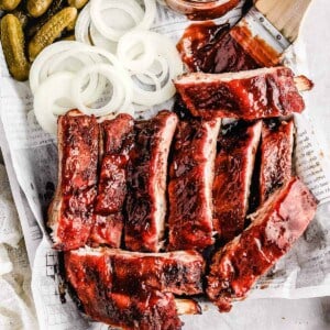 an overhead shot of traeger baby back ribs on a tray next to sliced white onions, dill pickles, and a jar of bbq sauce with a wooden brush