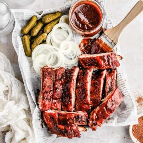 an over head shot of traeger baby back ribs, pickles, white onions, and a jar of bbq sauce with a wooden brush next to it.