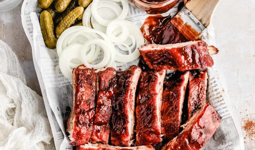 an over head shot of traeger baby back ribs, pickles, white onions, and a jar of bbq sauce with a wooden brush next to it.
