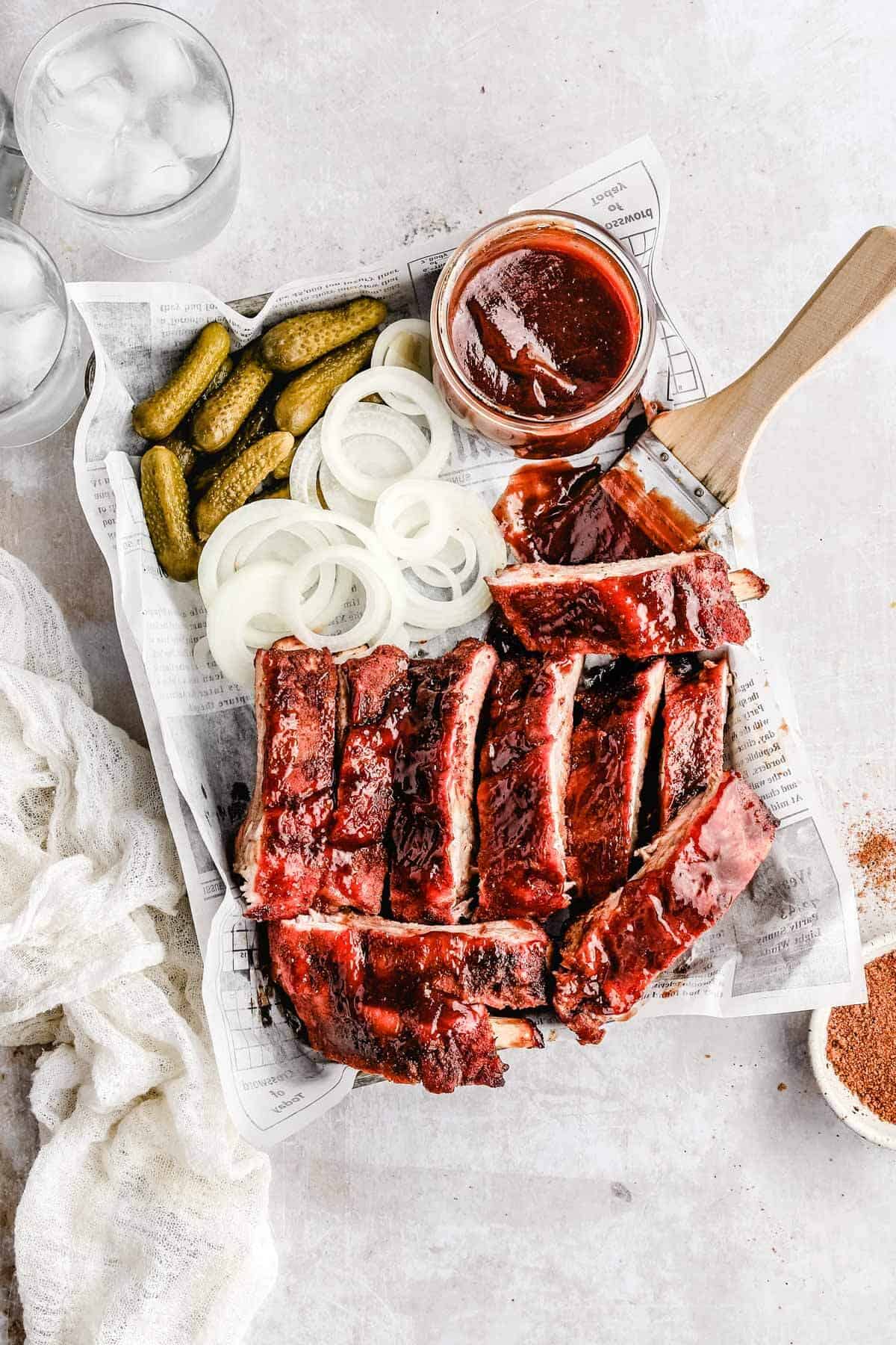 an over head shot of traeger baby back ribs, pickles, white onions, and a jar of bbq sauce with a wooden brush next to it. 
