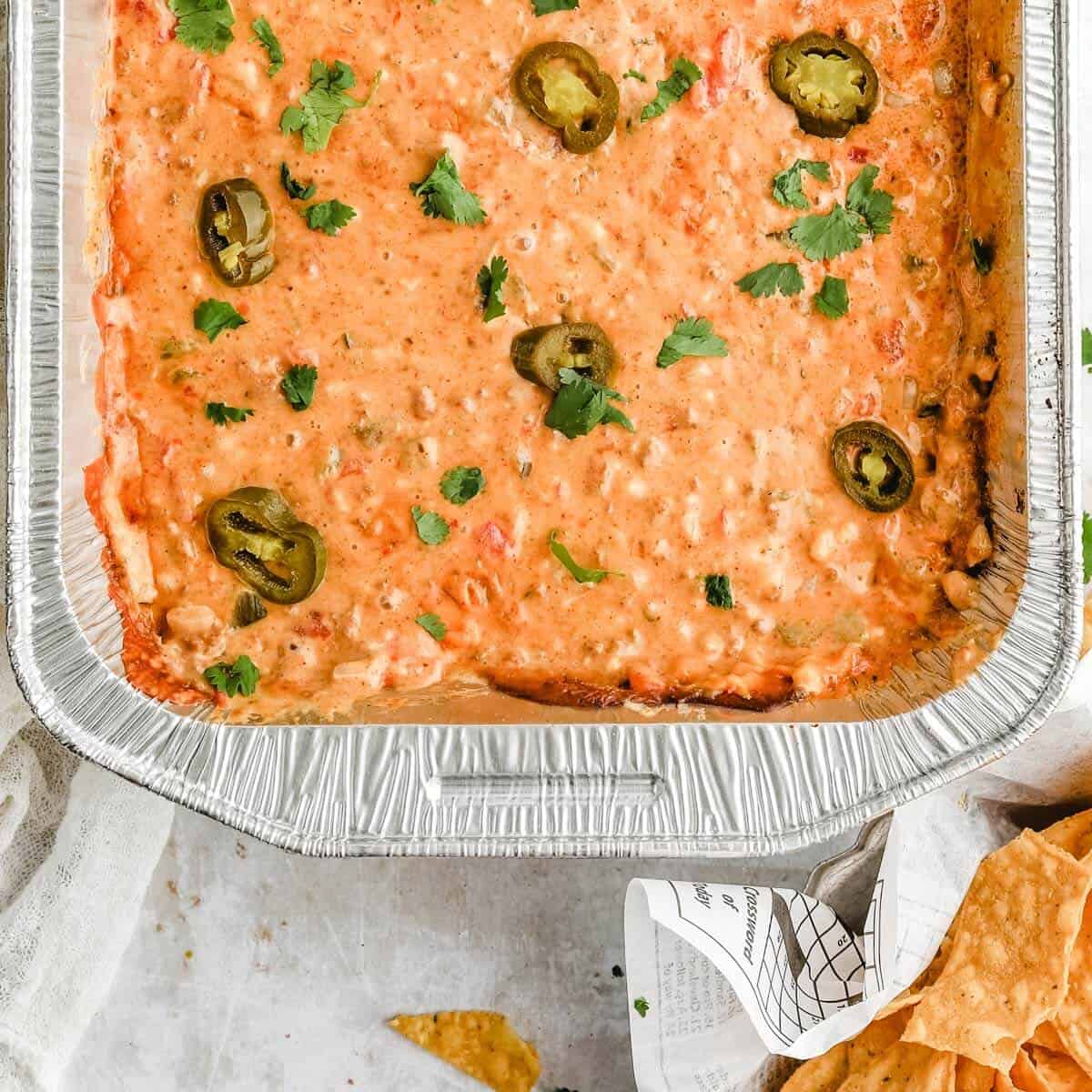 a photo of half a foil pan full of smoked queso dip topped with chopped cilantro and jalapeno slices next to a basket of tortilla chips