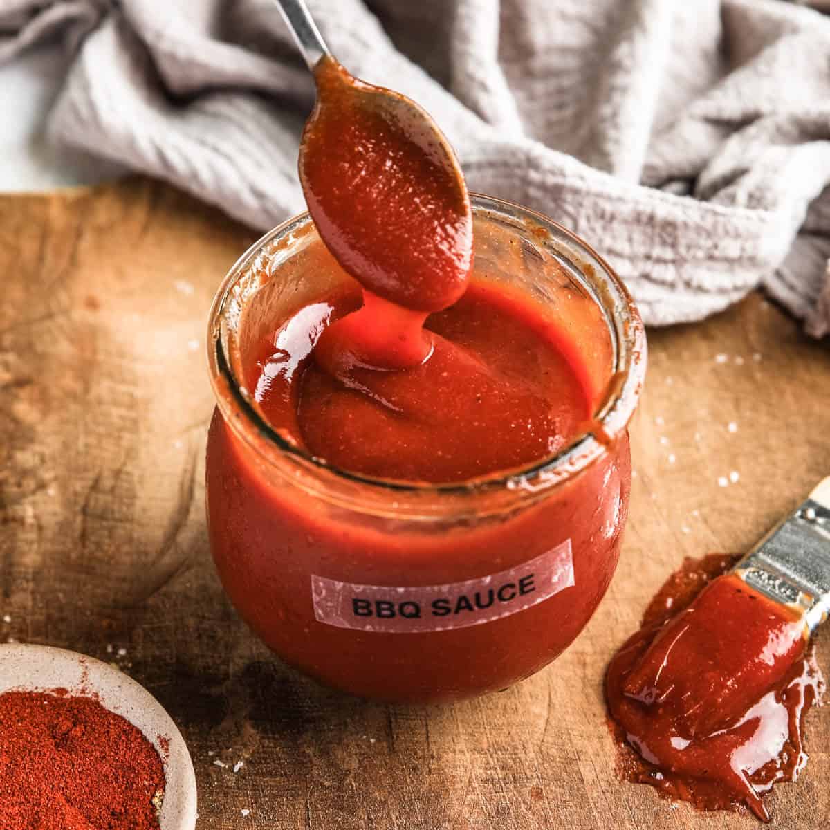 an overhead view of bbq sauce in a small glass jar with a silver spoon lifted above the glass to show the consistency of the bbq sauce.