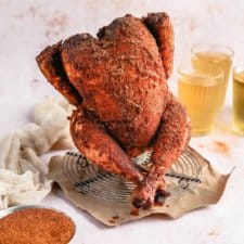 a closeup photo of a cooked Traeger beer can chicken covered in crispy dry rub on a wire rack next to a bowl of spices and a glass of beer