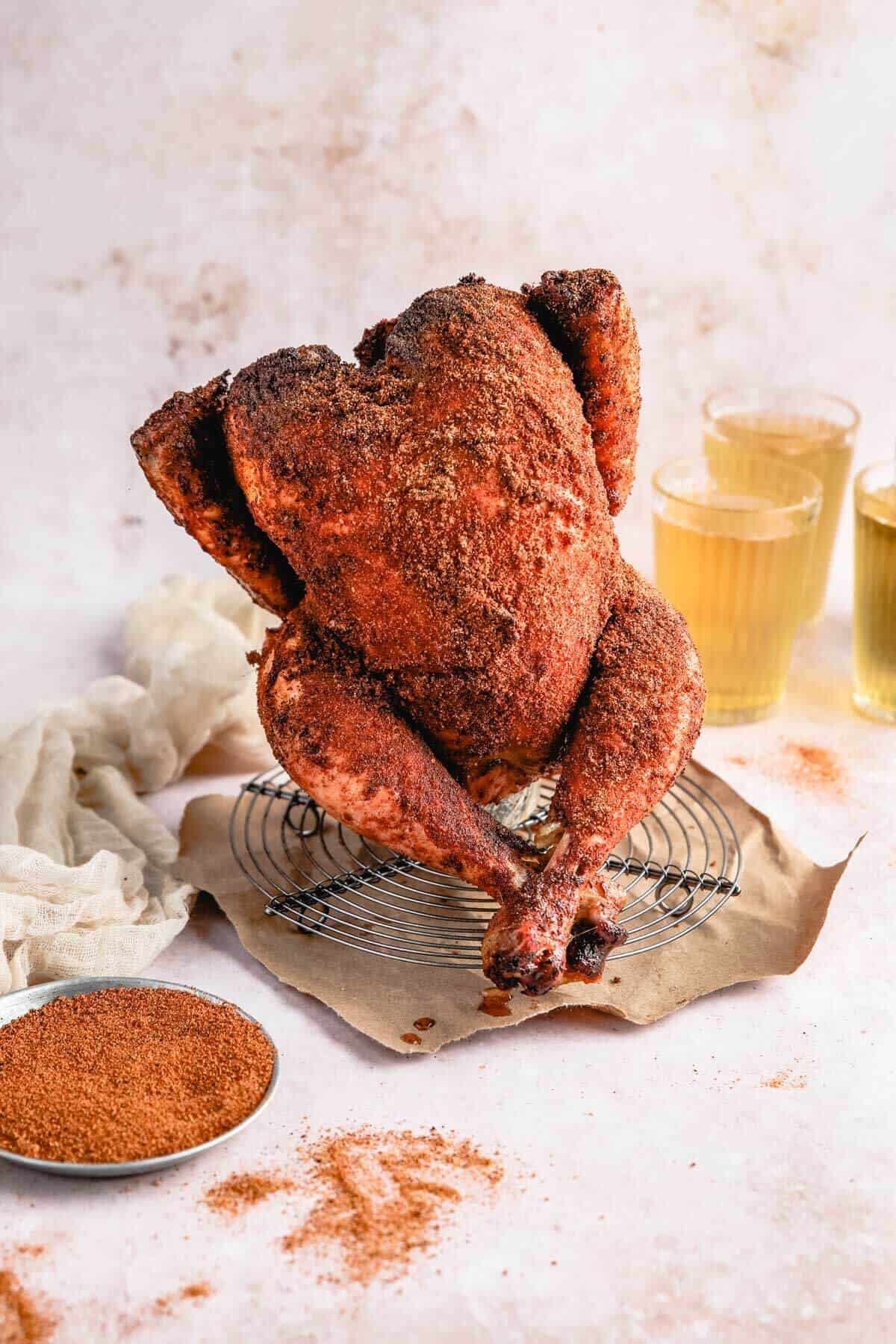 a whole smoked beer can chicken with homemade rub standing upright on a wire wrack net to a glass of beer