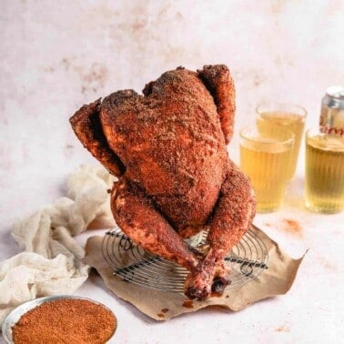 a closeup photo of a cooked Traeger beer can chicken covered in crispy dry rub on a wire rack next to a bowl of spices and a glass of beer