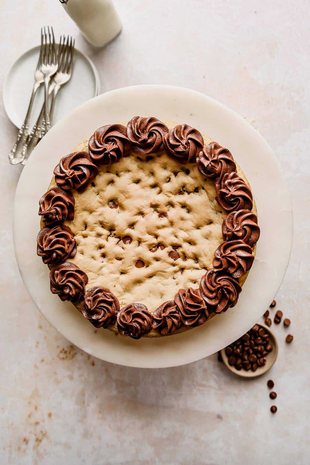 a photo of a whole cookie dough cake piped with chocolate buttercream frosting on a white plate with four silver forks resting on a plate next to it