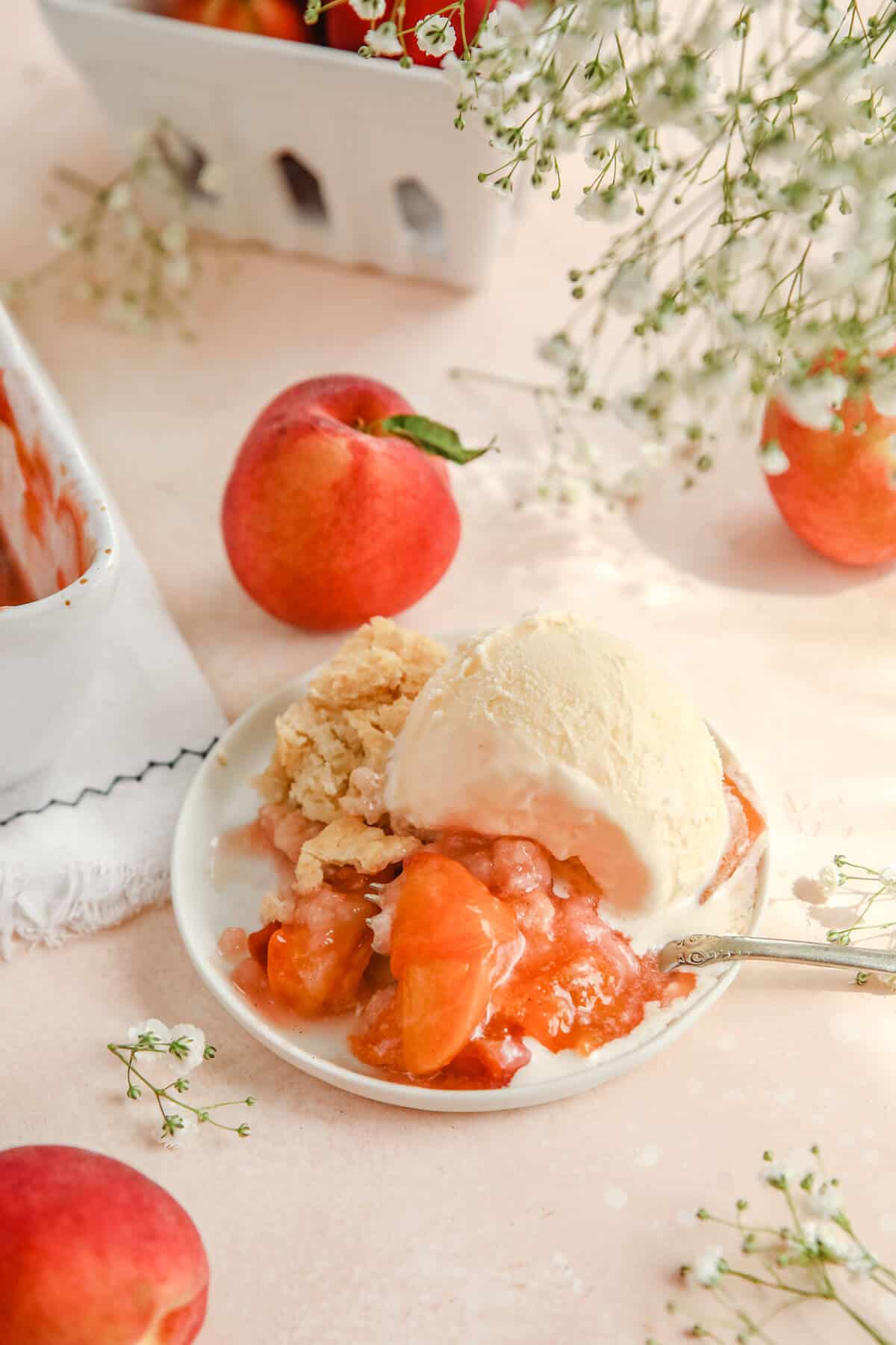 a slice of gluten-free peach cobbler on a white plate topped with vanilla ice cream with a silver fork resting next to it. 