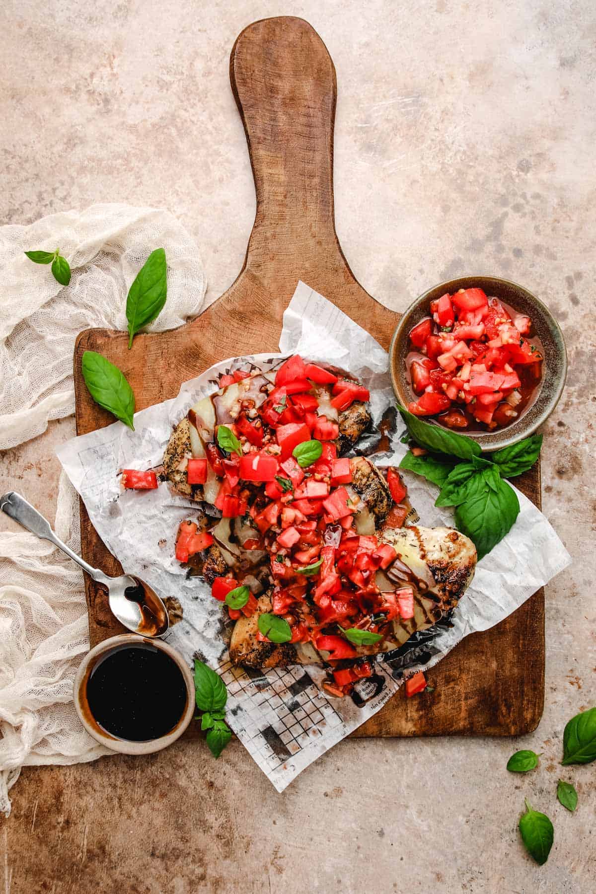 a wooden cutting board topped with three grilled bruschetta chicken breasts next to a bowl of tomatoes, balsamic vinegar, and fresh basil