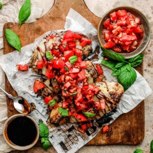 a wooden cutting board topped with three grilled bruschetta chicken breasts next to a bowl of tomatoes, balsamic vinegar, and fresh basil