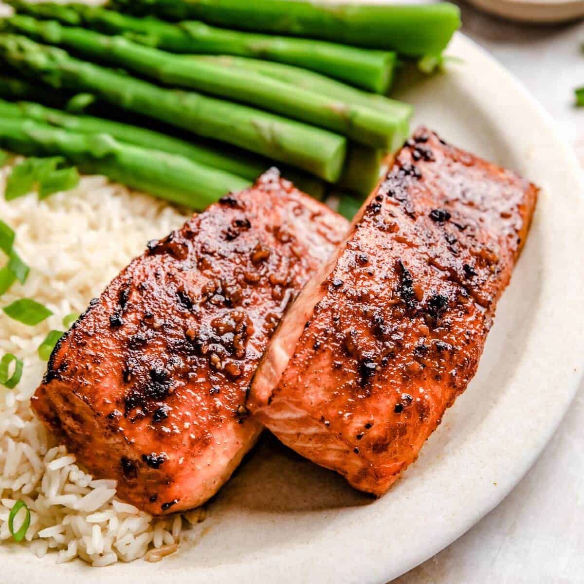 two honey garlic salmon filets on a white plate next to asparagus and rice.