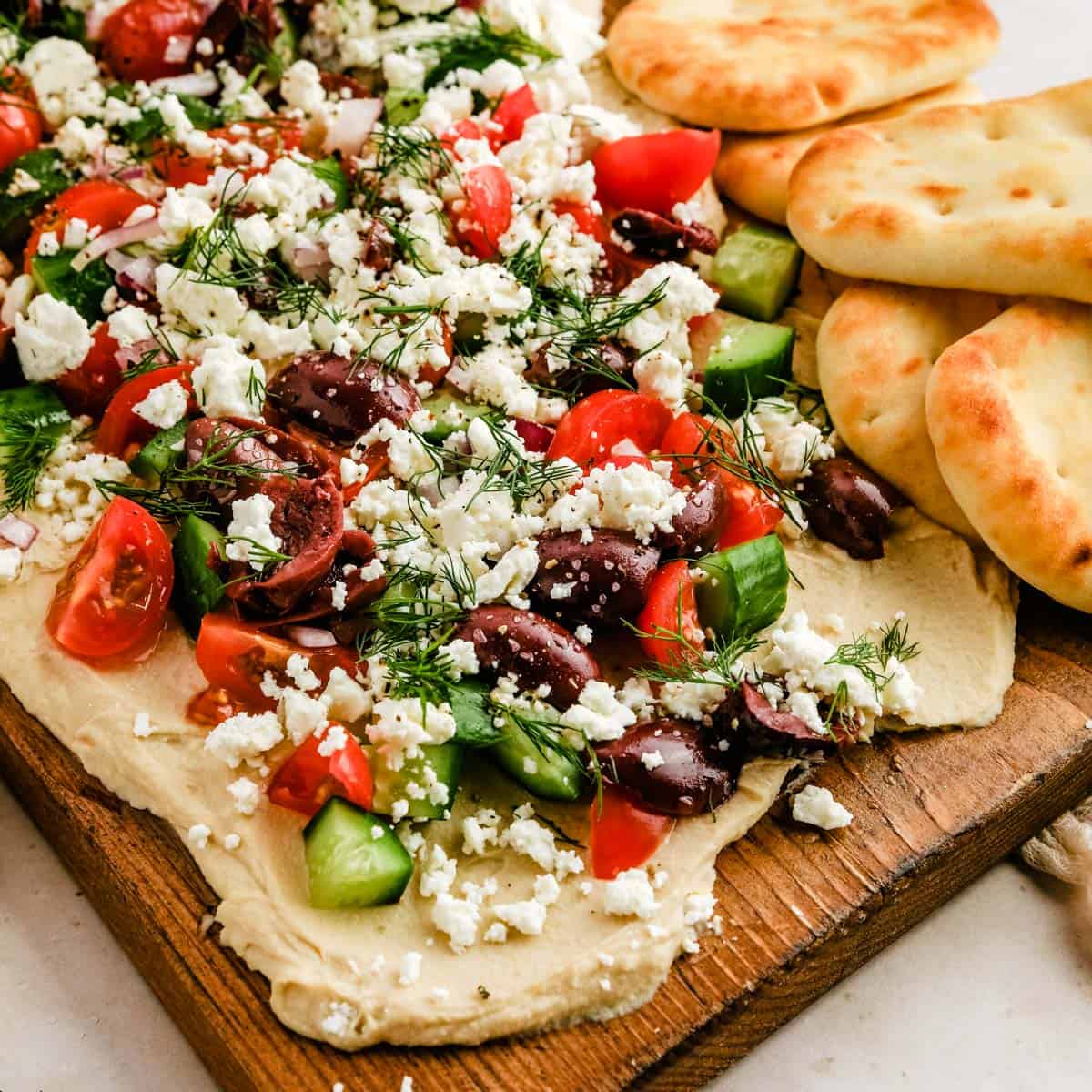 a close up of a cutting board with hummus, tomatoes, cucumbers, olives, and feta next to mini naan bread.