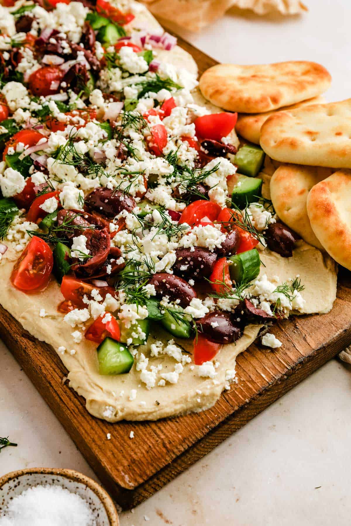 a close up of a cutting board with hummus, tomatoes, cucumbers, olives, and feta next to mini naan bread. 