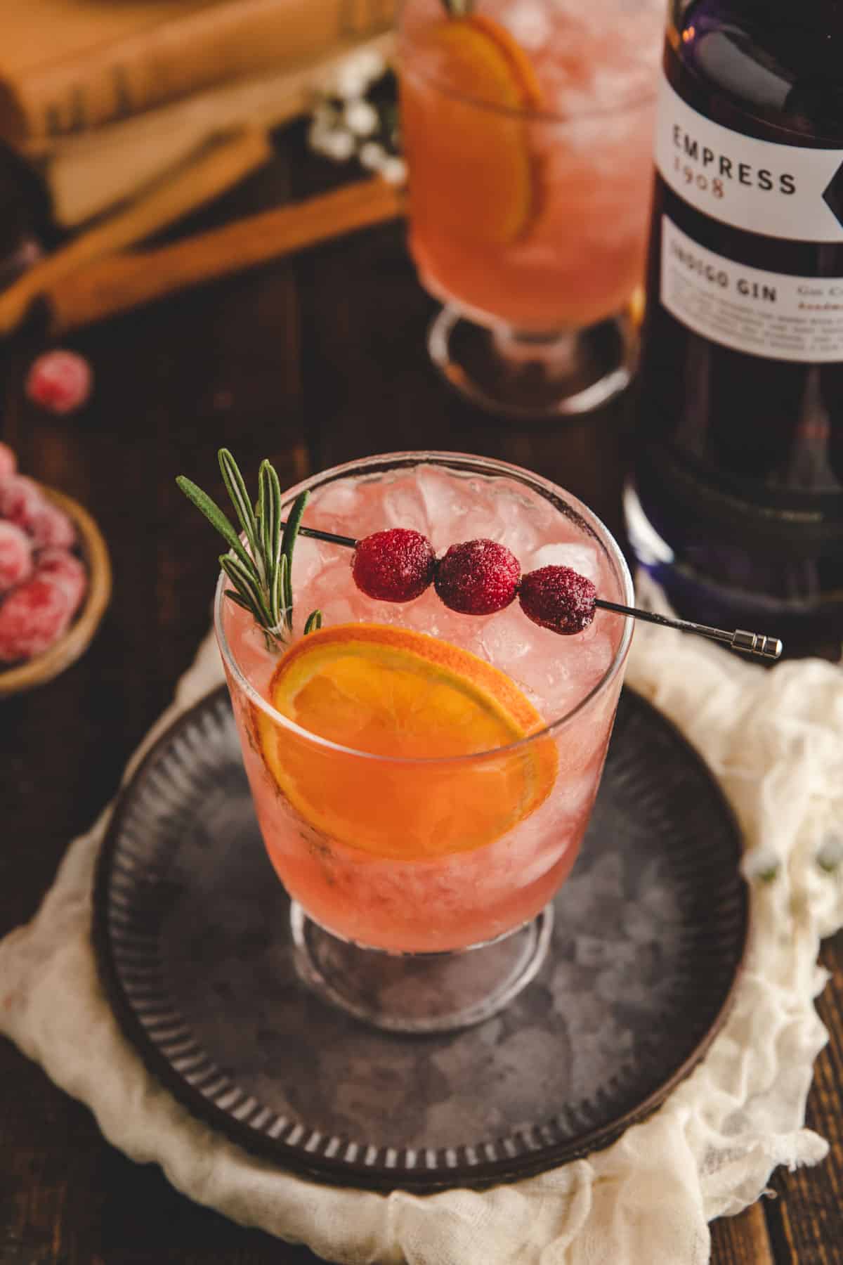a glass filled with cranberry gin cocktail and garnished with a sprig of rosemary, a dried orange, and candied cranberries