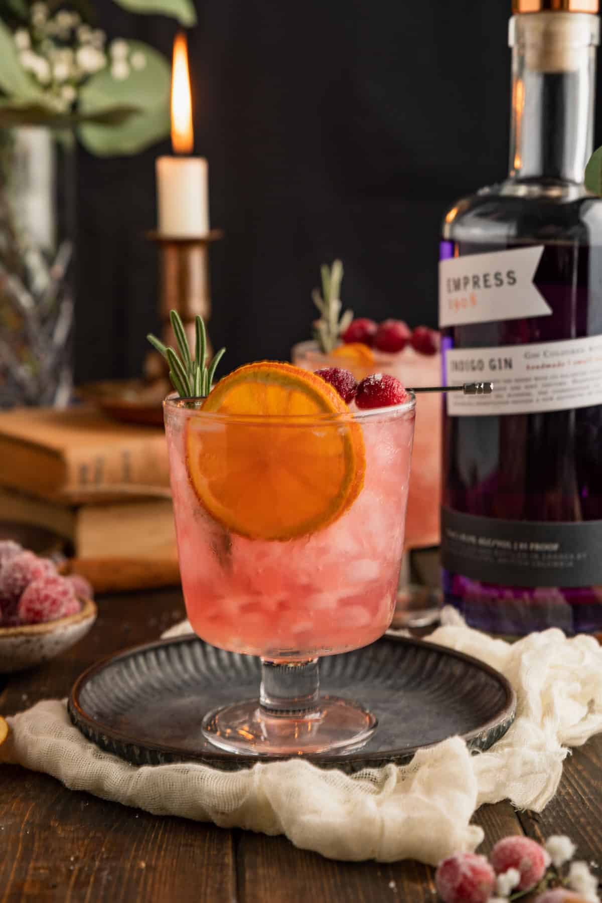 a closeup shot of a cranberry gin cocktail with a dried orange, rosemary and candied cranberries next to a bottle of gin