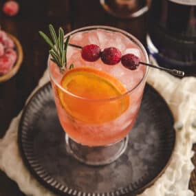 an overhead shot of a glass filled with cranberry gin cocktail and garnished with a sprig of rosemary, a dried orange, and candied cranberries