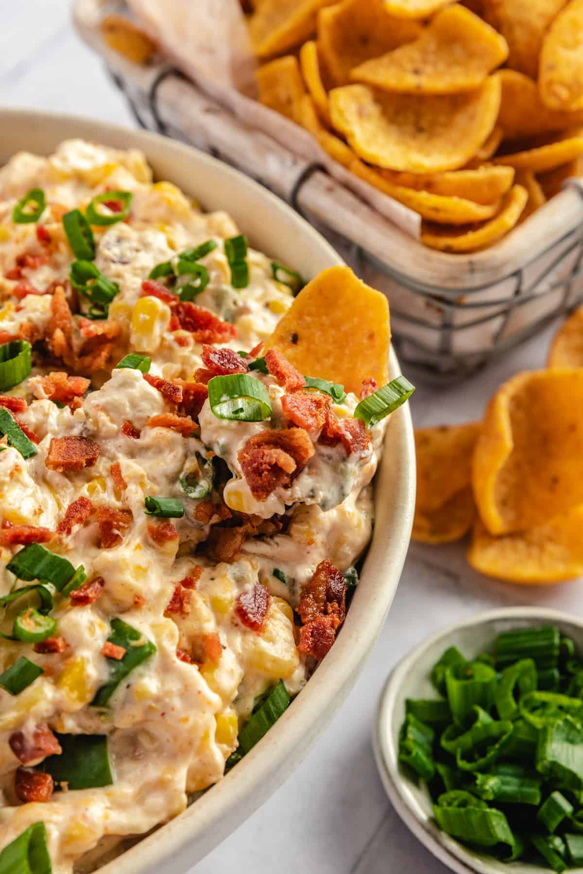 a closeup photo of a creamy, cheesy dip topped with bacon and green onion, next to a pile of corn chips.