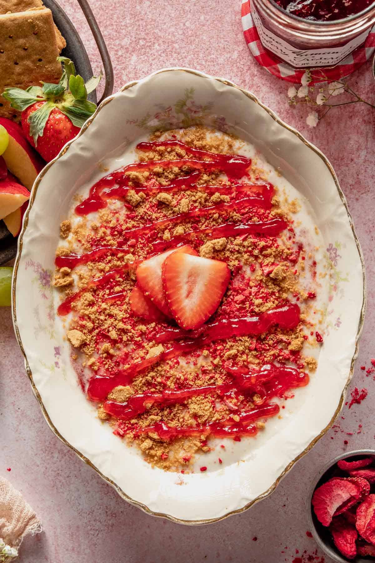 a bowl of strawberry dip topped with crumbled graham crackers, strawberry jam, and fresh strawberry slices. 