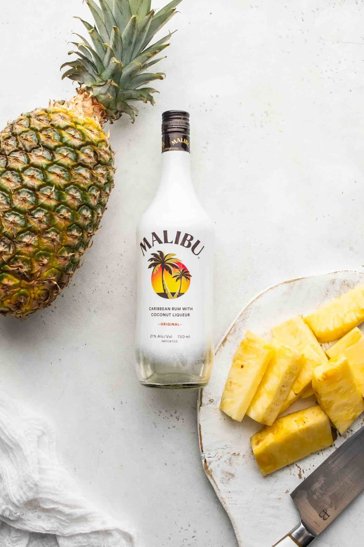 a large pineapple, bottle of malibu rum, and a plate of pineapple spears
