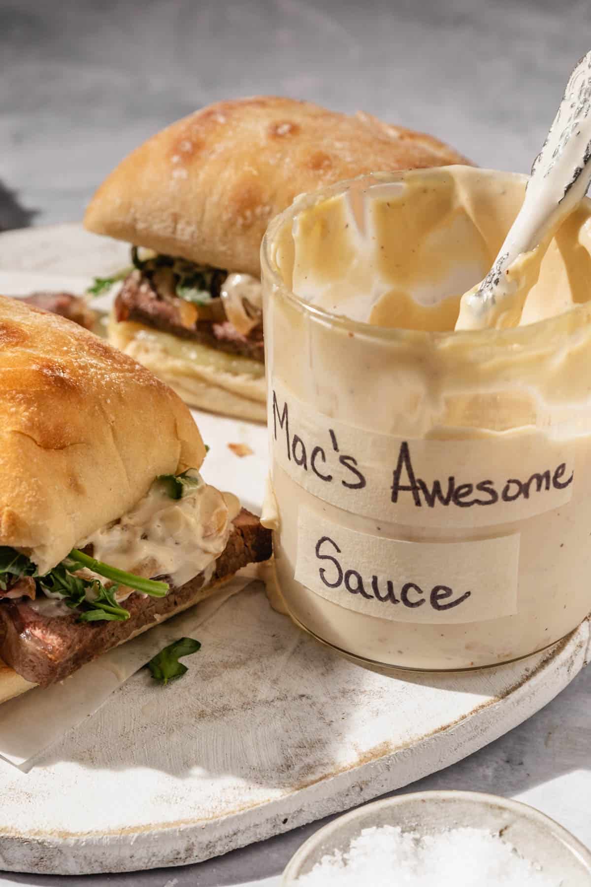 an up close photo of a jar of mac's awesome sauce next to a sandwich. 