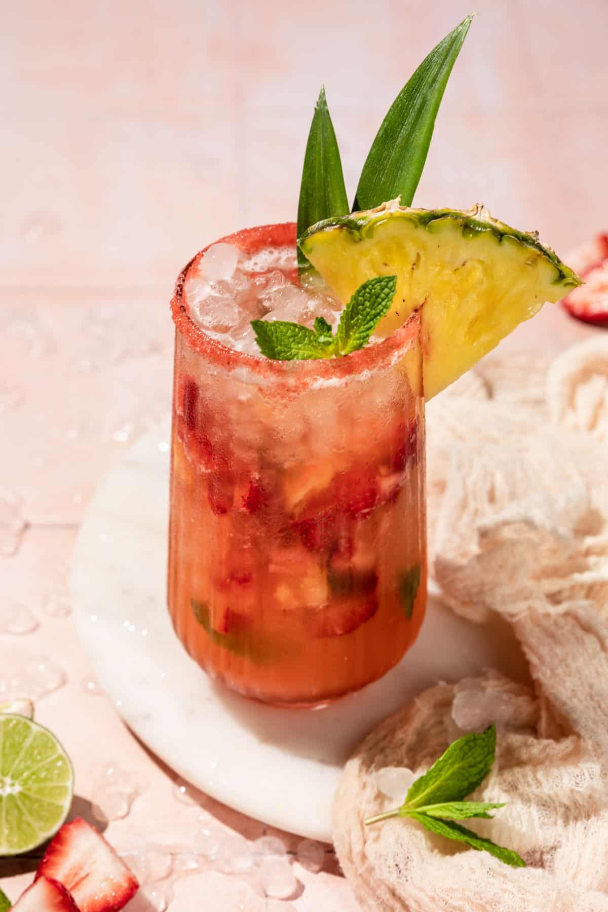 a tall glass filled with strawberry pineapple mojito mocktail and garnished with a mint sprig and pineapple wedge. 