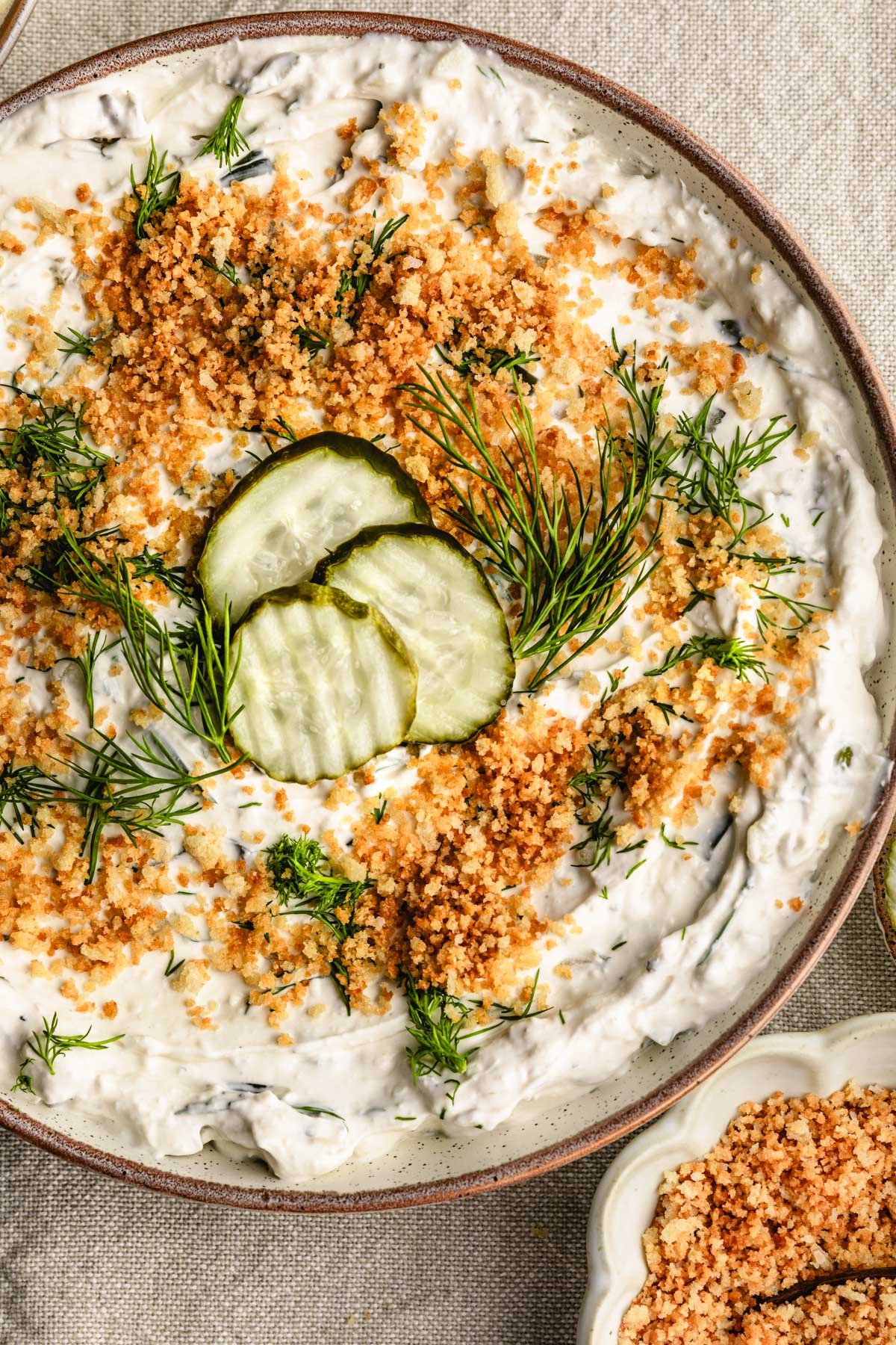 a bowl of fried pickle dip topped with three dill pickles, fresh herbs, and toasted Panko bread crumbs