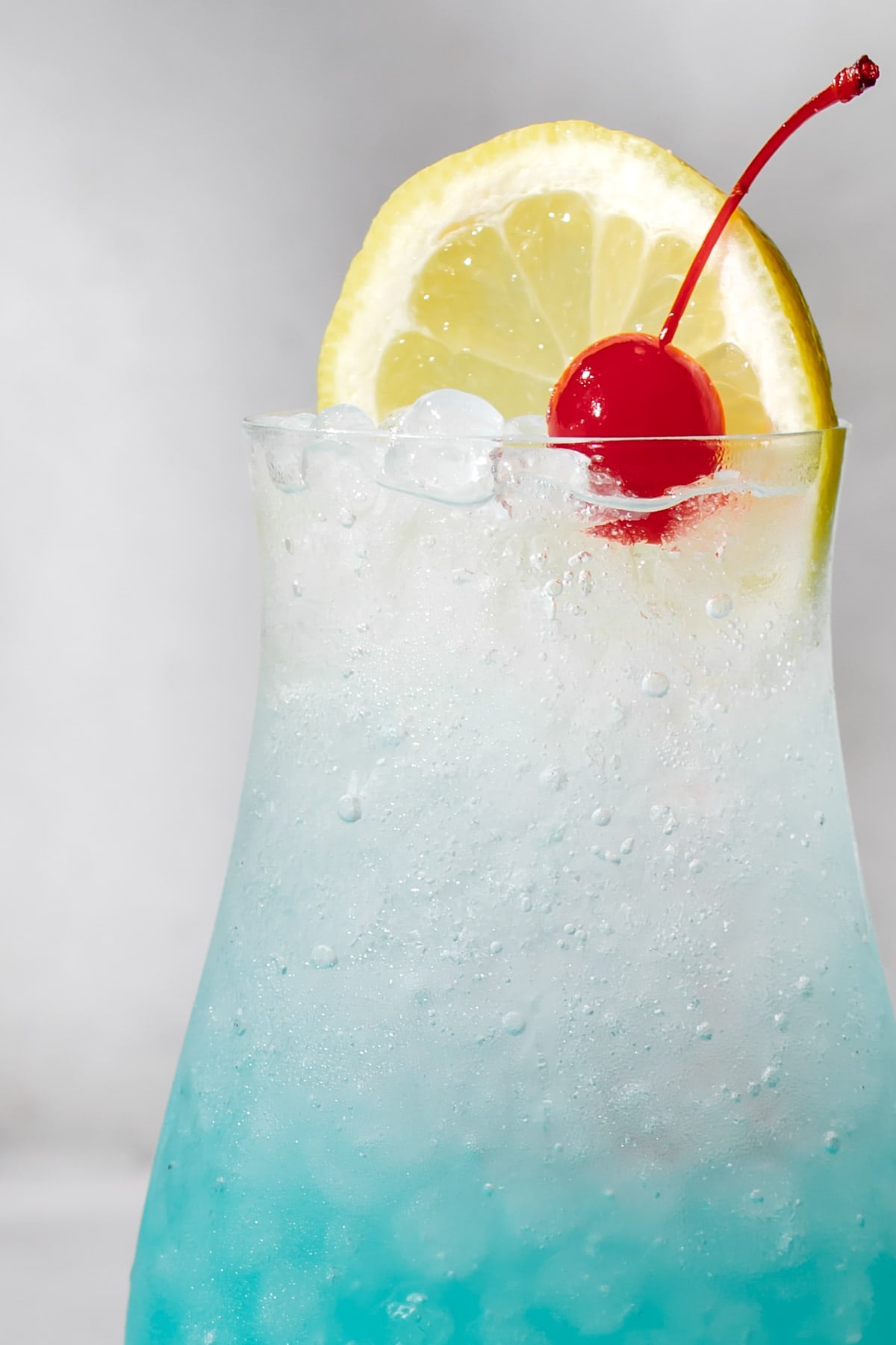 a close up shot showing the top of a hurricane glass filled with sparkling blue lagoon mocktail and topped with a lemon and a cherry