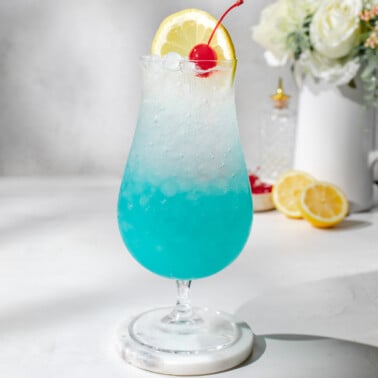 a hurricane glass filled with a blue lagoon mocktail and garnished with a maraschino cherry and a lemon slice.
