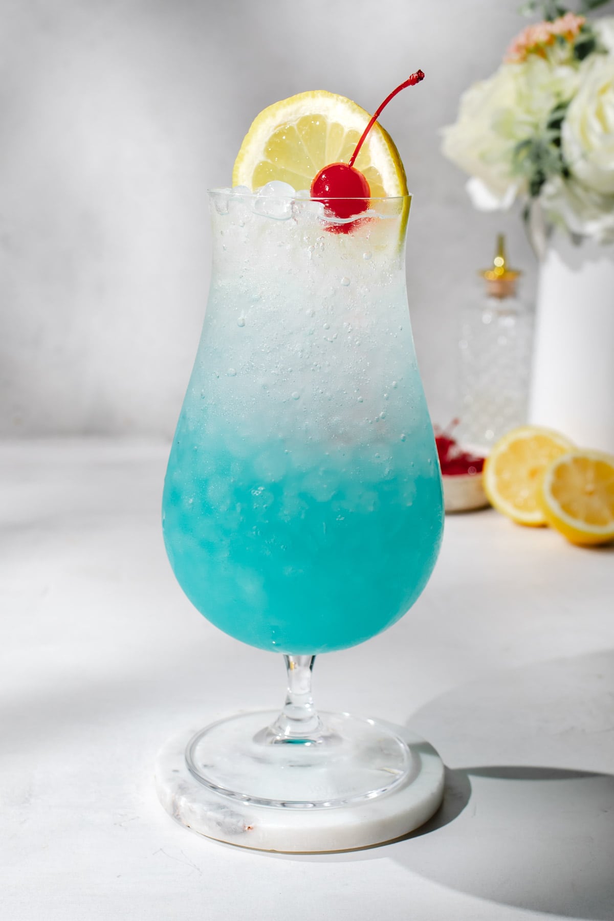 a hurricane glass filled with a blue lagoon mocktail and garnished with a maraschino cherry and a lemon slice. 