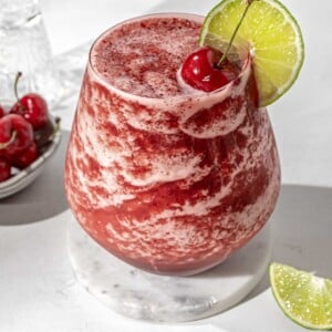 a vodka cherry limeade slushy in a glass garnished with cherries and lime.