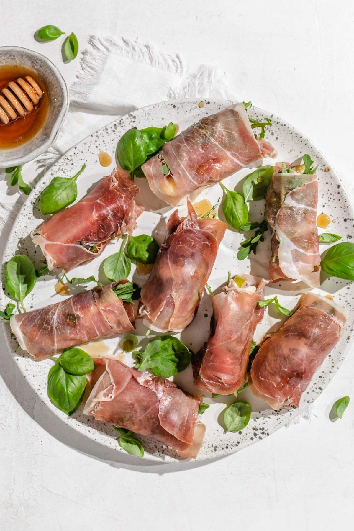 an overhead shot of 8 prosciutto wrapped mozzarella roll ups on a speckled platter. 