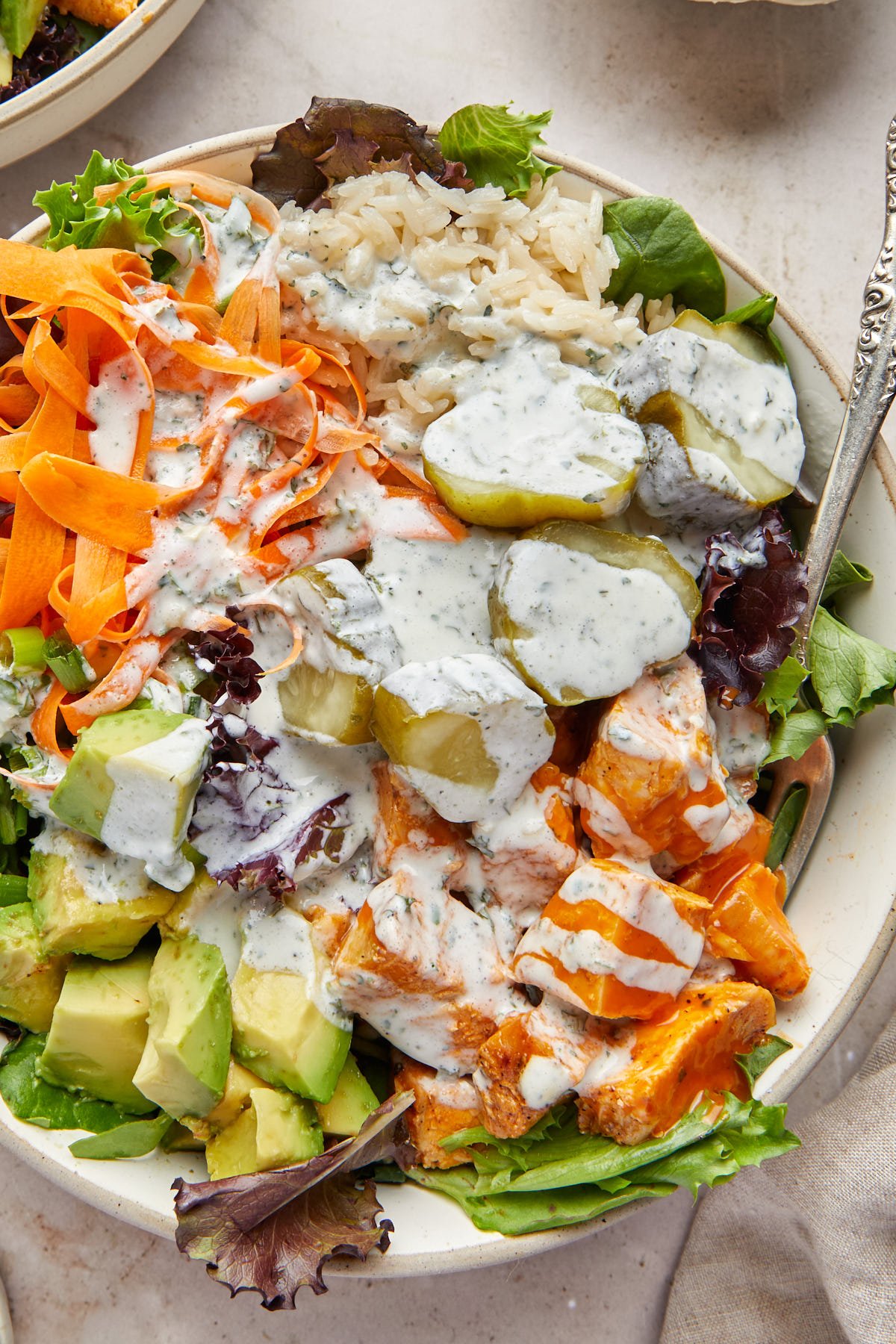 a bowl filled with buffalo chicken, pickles, avocado, carrot, greens, and drizzled with ranch. 