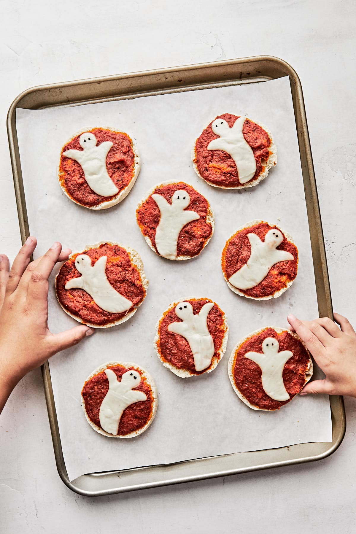 a baking sheet pan with parchment paper topped with ghost English muffin pizzas and two kid hands reaching in top grab them