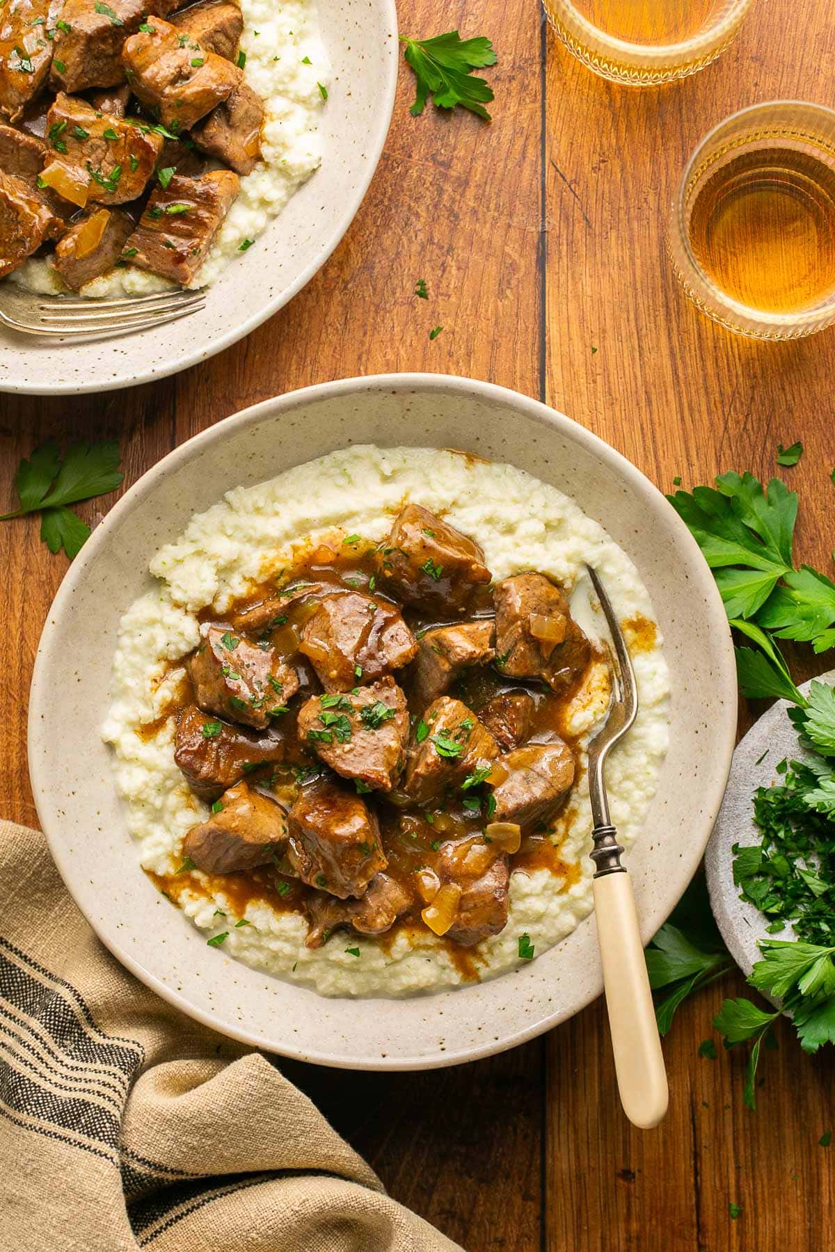 Beef Tips and Gravy over cauliflower mash in a cream bowl on a wood backdrop with chopped parsley 