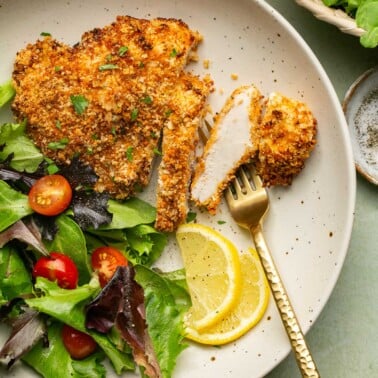 a gold fork resting next to a chicken cutlet with a side salad and lemon slices.