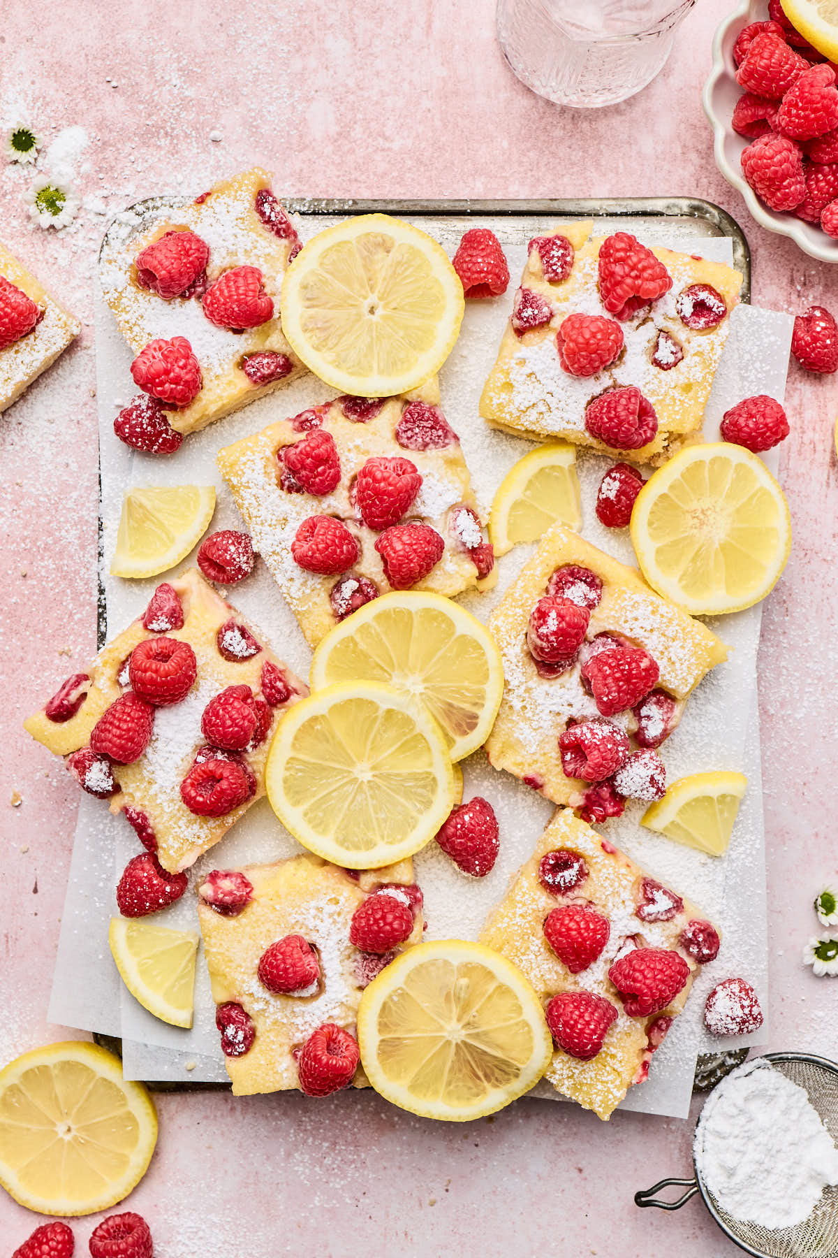 a sheet pan with raspberry lemon bars resting atop next to fresh raspberries and lemon slices