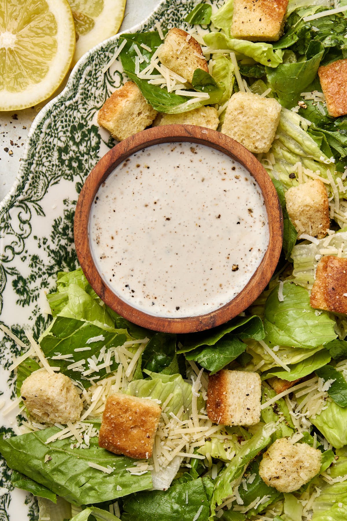 A Caesar salad with romaine lettuce, croutons, and grated parmesan is arranged on a decorative plate. A small wooden bowl filled with high protein caesar dressing is placed on top. Lemon slices are partially visible in the corner.