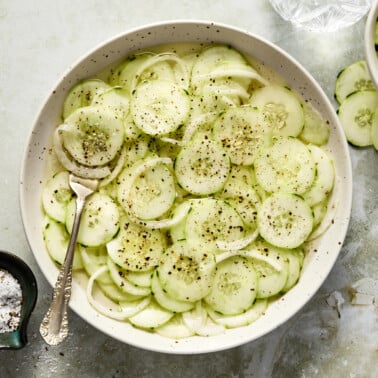 an overhead shot of sliced cucumbers in a white bowl with sliced onions and black pepper