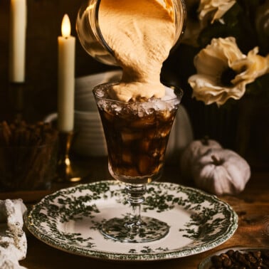 a hand pouring pumpkin cold foam onto a glass of iced coffee