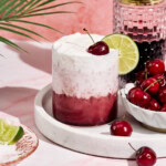 A layered cherry and cream drink in a glass topped with whipped cream, a lime slice, and a cherry, sits on a marble tray. Beside it, a bowl of cherries and a tall, patterned pink glass. A palm leaf casts a shadow on a pink backdrop.