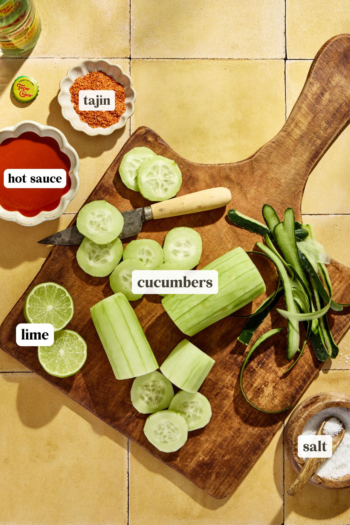 recipe ingredients on a wooden cutting board and labeled, hot sauce, lime, cucumbers, tajin, and salt.