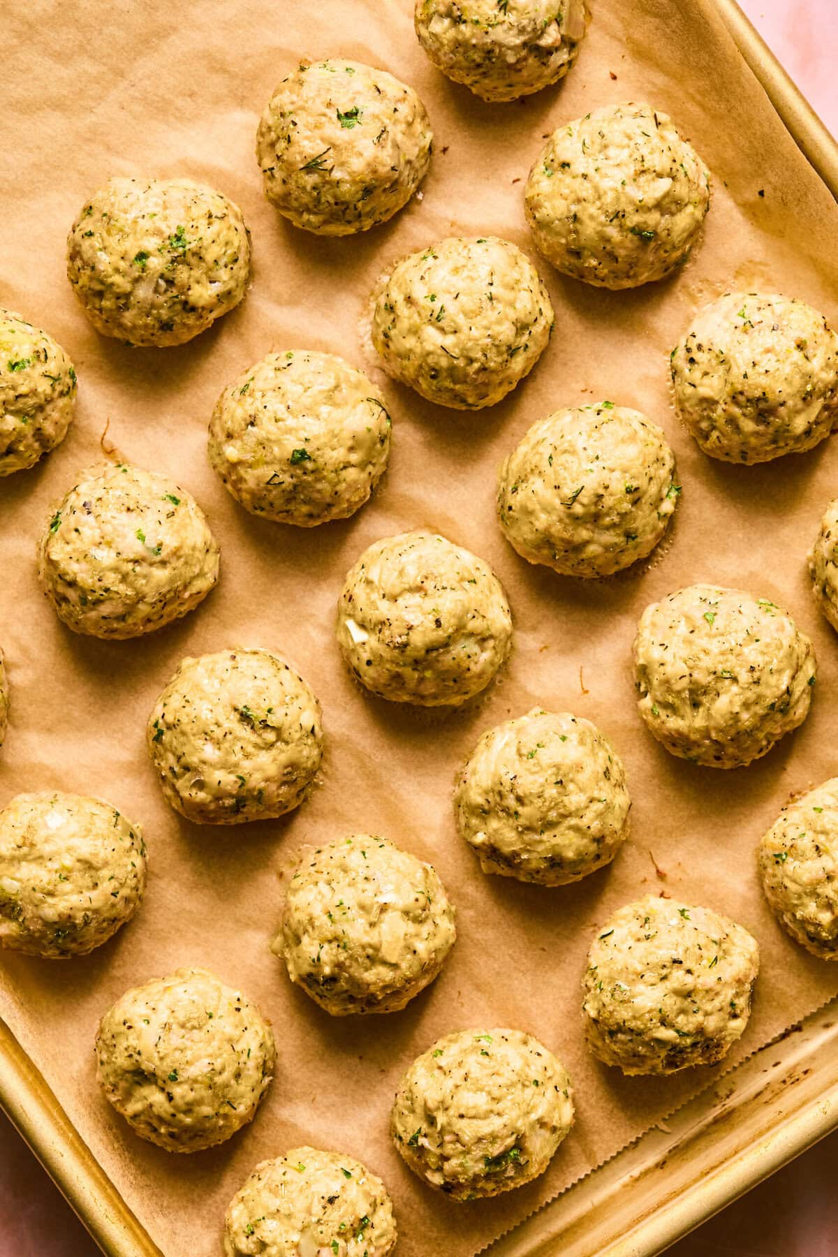 Rows of uniformly shaped, uncooked Greek chicken meatballs are arranged on a parchment-lined baking sheet. The balls are speckled with herbs and spices, giving them a textured appearance.