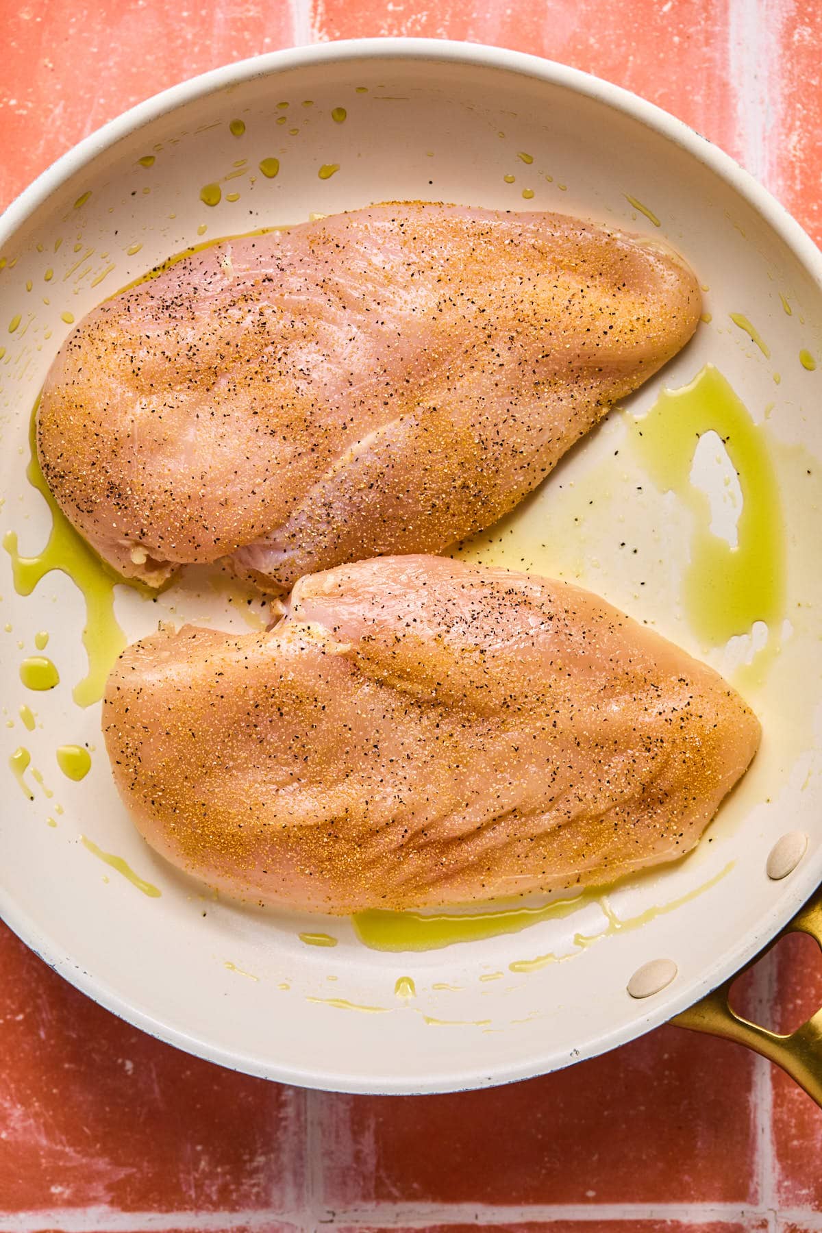 Raw chicken breasts seasoned with pepper in a white skillet, drizzled with olive oil. The skillet is placed on a red and white tiled surface.